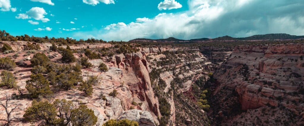 Panoramic view of a rugged canyon with steep rock walls, dotted with green shrubs. The sky is bright blue with scattered clouds, and theres a mix of sunlit and shadowed areas across the landscape.