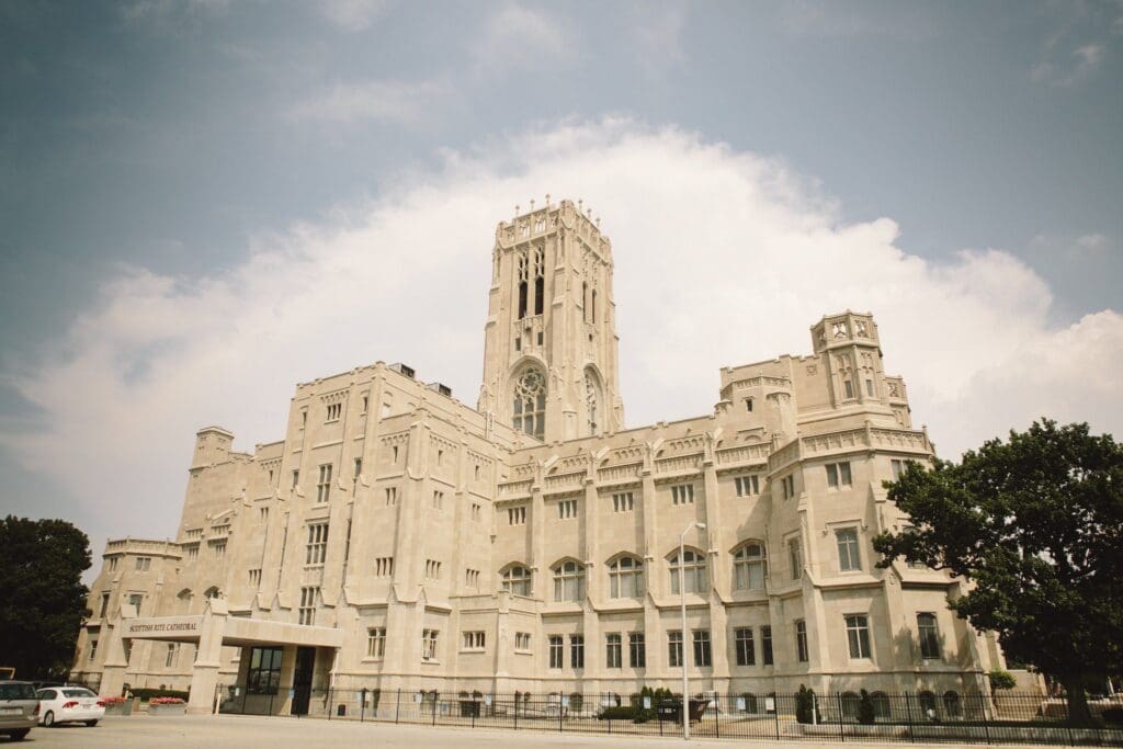 A grand, beige stone building with intricate architectural details and a tall central tower. The structure stands under a partly cloudy sky, with some trees and parked cars in the foreground.