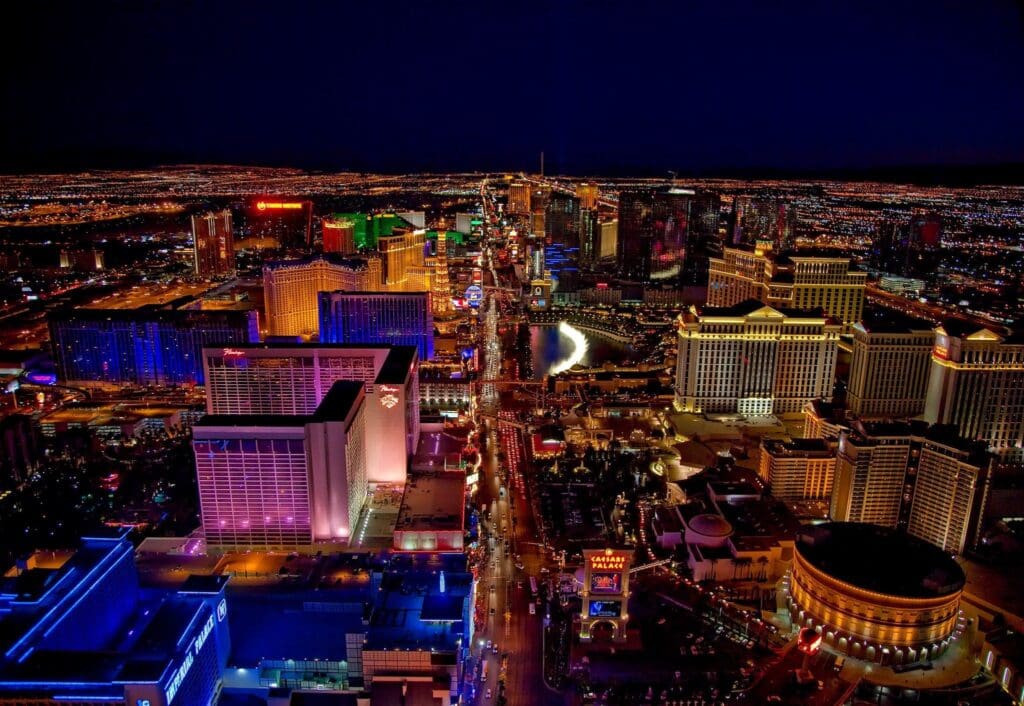 Aerial view of the vibrant Las Vegas Strip at night, illuminated by bright lights from casinos and hotels. The cityscape features iconic buildings and colorful signage, with a dark sky contrasting the vivid city lights.