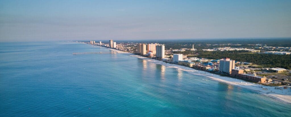 Aerial view of a coastal city with tall beachfront buildings lining a long stretch of sandy beach. The blue ocean extends to the horizon, and a pier is visible. The skyline includes both urban and green areas under a clear sky.