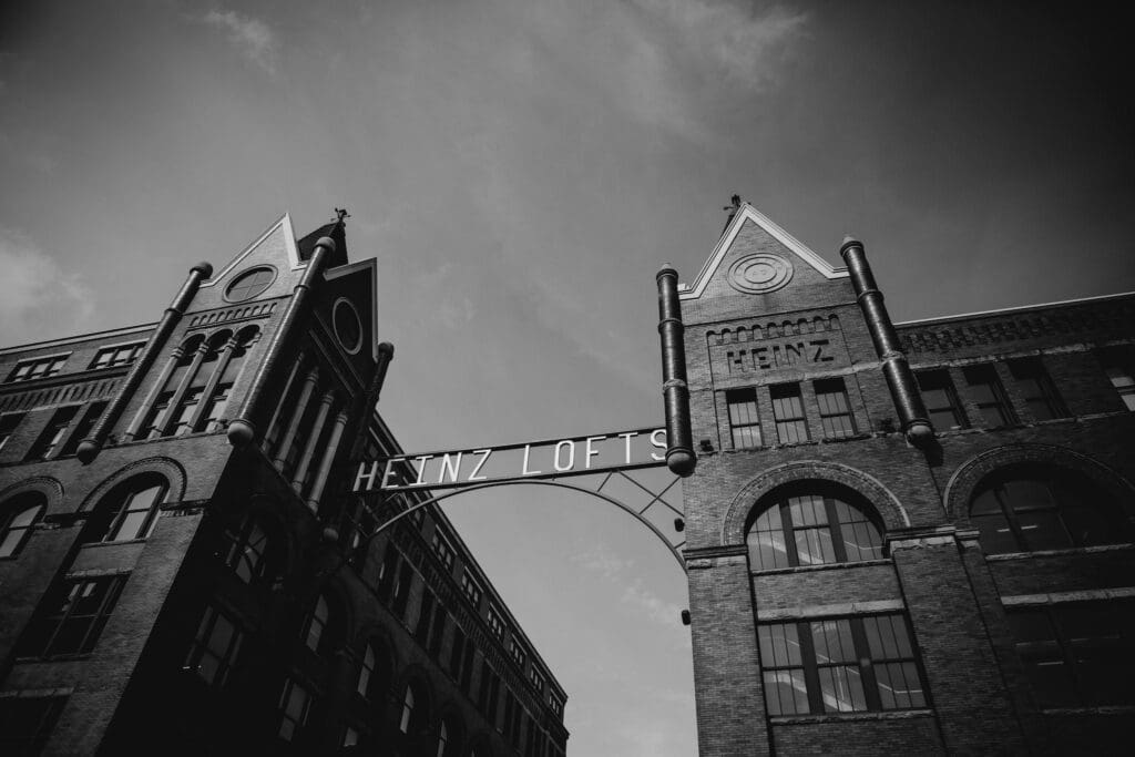 A black and white image of historic brick buildings with pointed roofs, connected by an archway sign reading Heinz Lofts. The architectural details include large arched windows and decorative brickwork. The sky is partially cloudy.