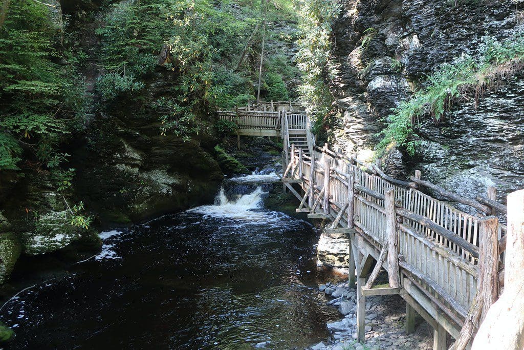 Wooden walkways run alongside a narrow, dark river with small waterfalls, surrounded by rocky cliffs and lush green foliage. Sunlight filters through the trees, illuminating parts of the scene.