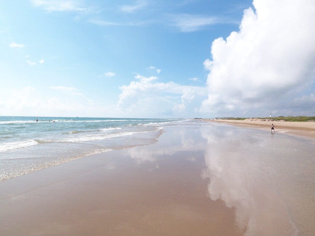 A tranquil beach scene with gentle waves lapping the sandy shore under a clear blue sky. White clouds are reflected on the wet sand. In the distance, a few people stroll along the beach enjoying the calm setting.