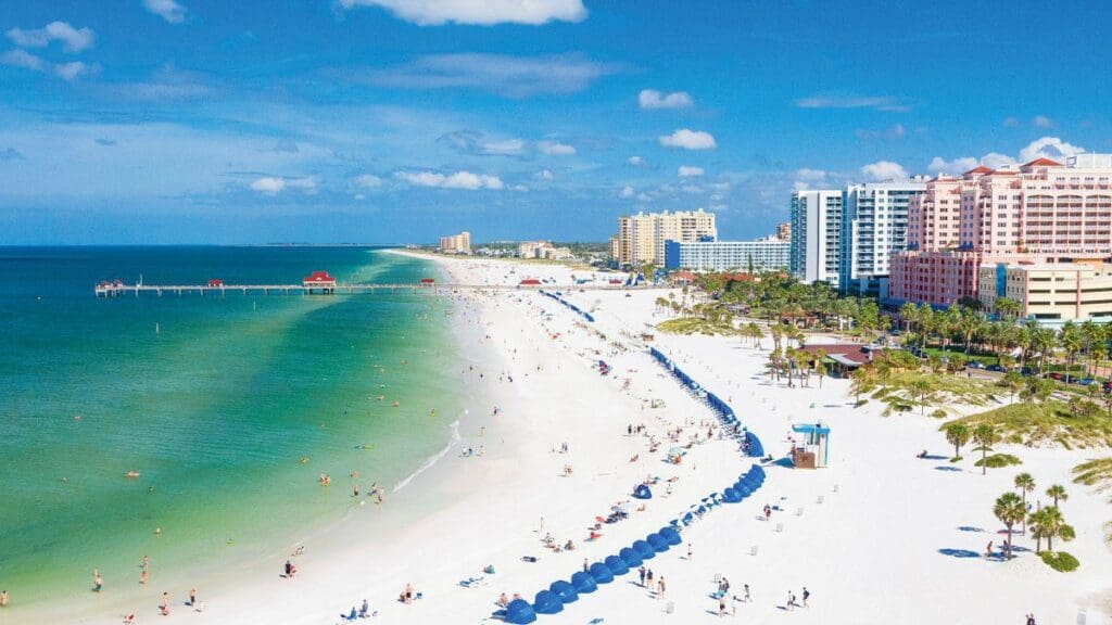 Aerial view of a beach with white sand and turquoise water. People relax under blue umbrellas. Nearby are tall buildings and a pier extending into the water. The sky is partly cloudy.