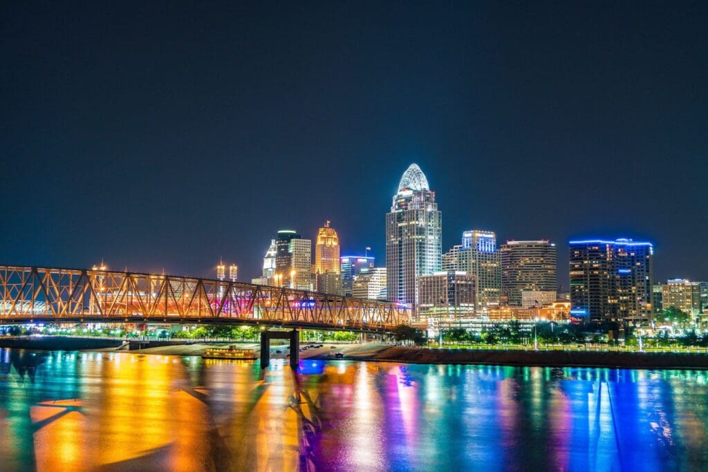 A vibrant cityscape at night featuring a brightly lit skyline with tall buildings and a bridge in the foreground. The reflection of colorful lights dances on the calm river below.