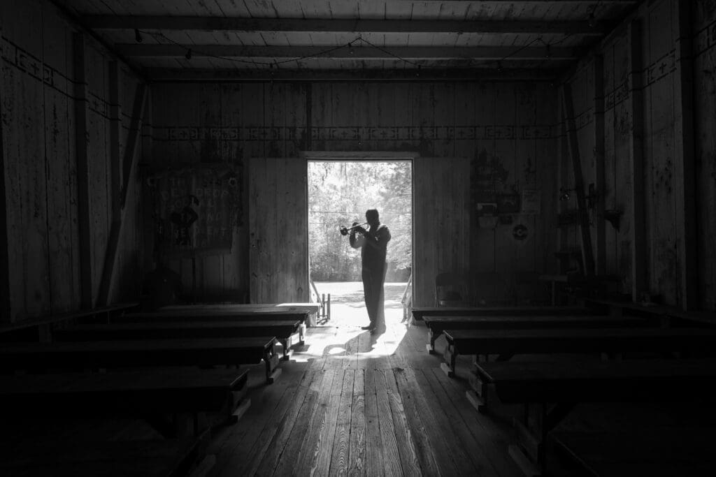 A silhouette of a person playing the trumpet stands in a sunlit doorway inside a rustic wooden room, with benches lined up on either side. The light creates a dramatic contrast, highlighting the musician against the bright outdoors.