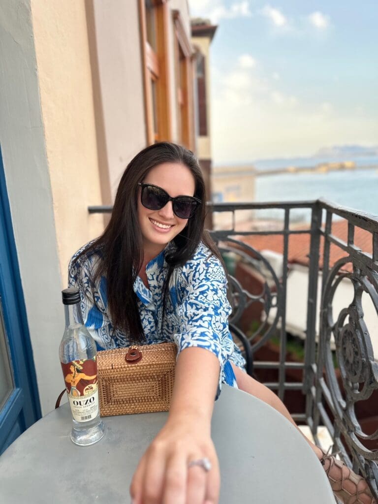 A woman with long dark hair wearing sunglasses and a blue and white patterned dress sits at a balcony table. She holds out her hand, displaying a ring. A bottle and a woven bag are on the table. The background shows a seaside view and rooftops.