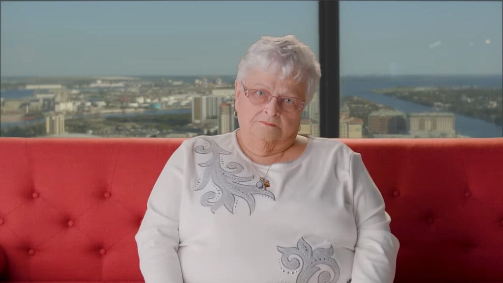 An older woman with short gray hair and glasses sits on a red couch. She is wearing a white top with decorative patterns. Behind her is a large window showing a cityscape with water and buildings under a clear sky.