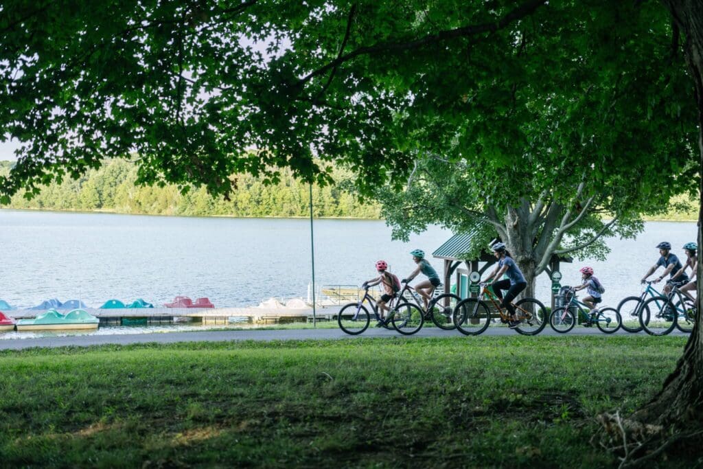 A group of people wearing helmets are riding bicycles on a path beside a lake. Paddle boats are visible on the water, and lush green trees provide shade. It is a sunny day with a tranquil outdoor setting.