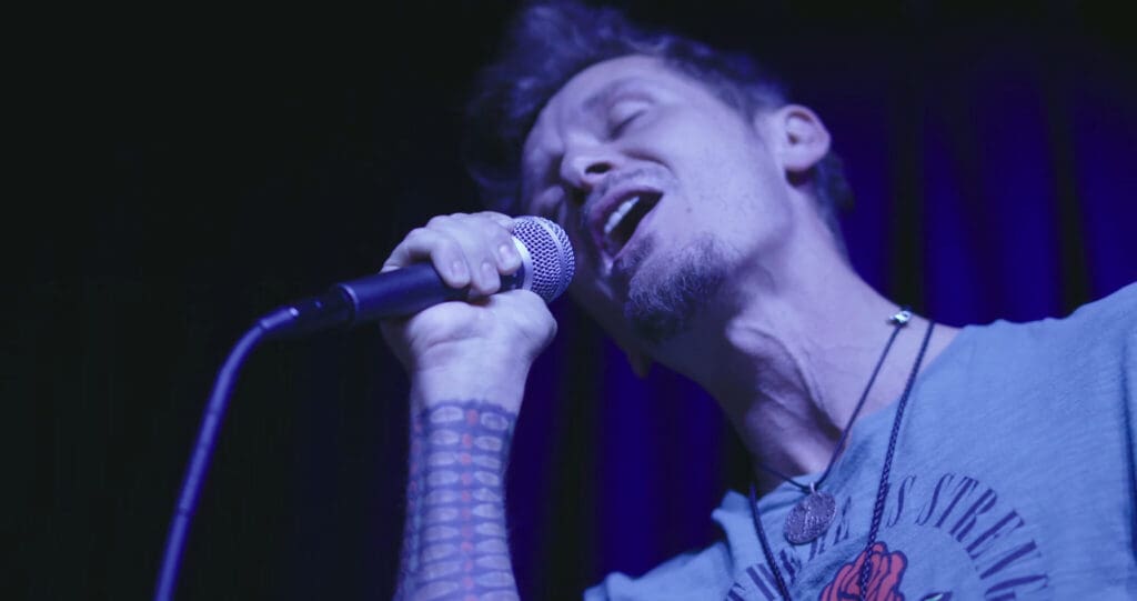 A man passionately sings into a microphone on stage. He has a tattooed arm and is wearing a gray shirt with a design. The lighting casts a blue hue on the scene, with curtains visible in the background.