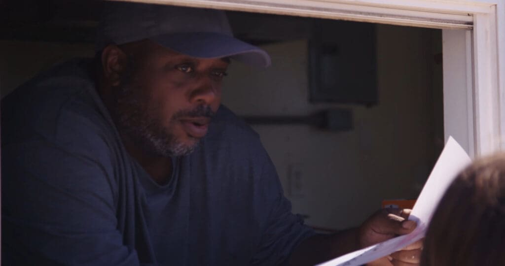 A person wearing a cap and a dark shirt looks out from a window, holding a piece of paper. The setting is dimly lit, suggesting an indoor or shaded area. Part of another persons head is visible in the foreground.