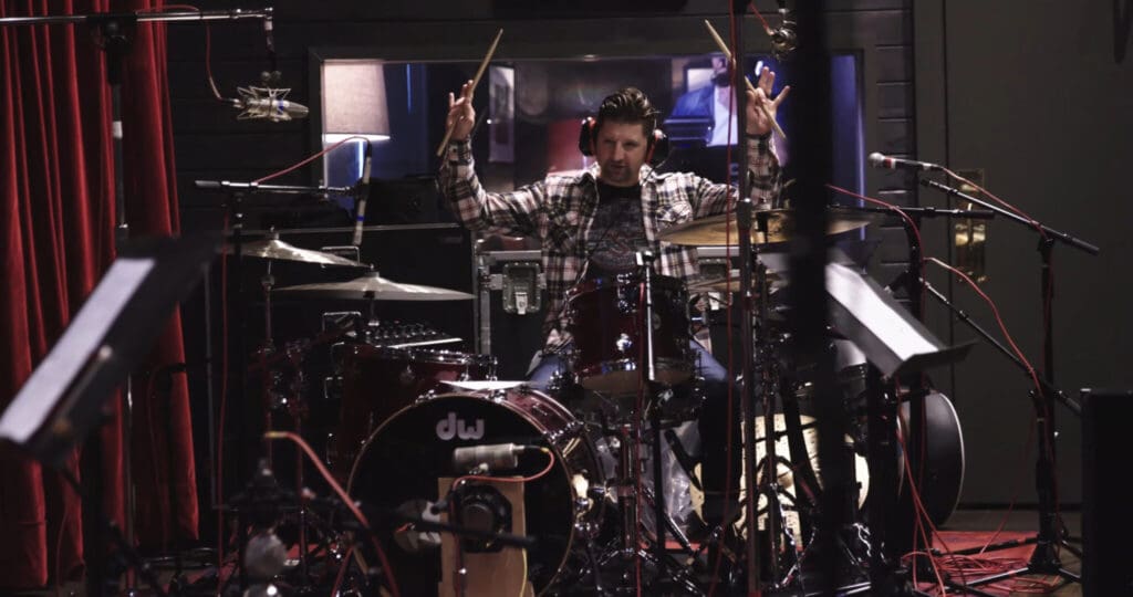 A drummer is energetically playing a drum set in a recording studio. He is wearing a plaid shirt and holding drumsticks in the air. The room is filled with various microphones and equipment, and a large window is visible behind him.