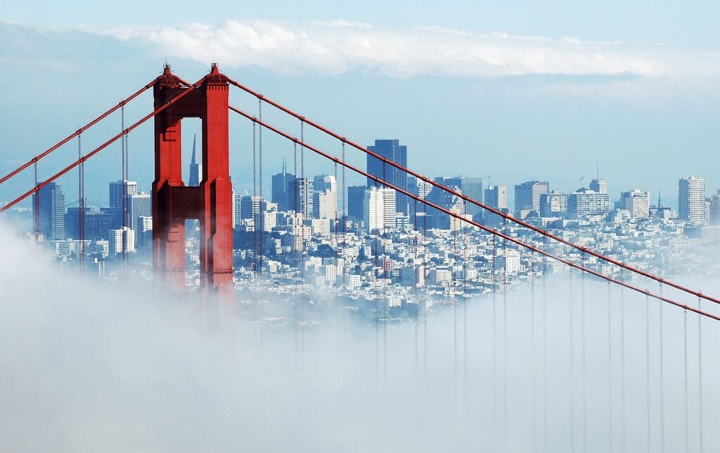 The top of the Golden Gate Bridge peeks through dense fog, with the skyline of San Francisco visible in the background under a clear blue sky.