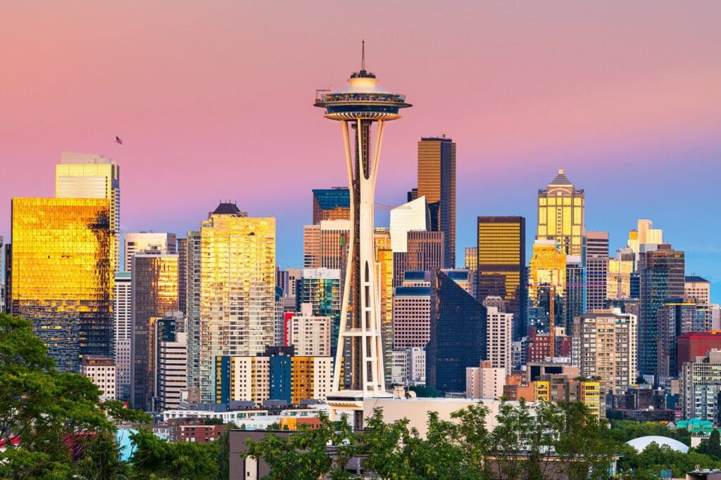 Seattle skyline at sunset with the Space Needle in the foreground. The sky is a gradient of pink, purple, and blue, and the buildings reflect a warm golden light. Trees are visible at the bottom of the image.
