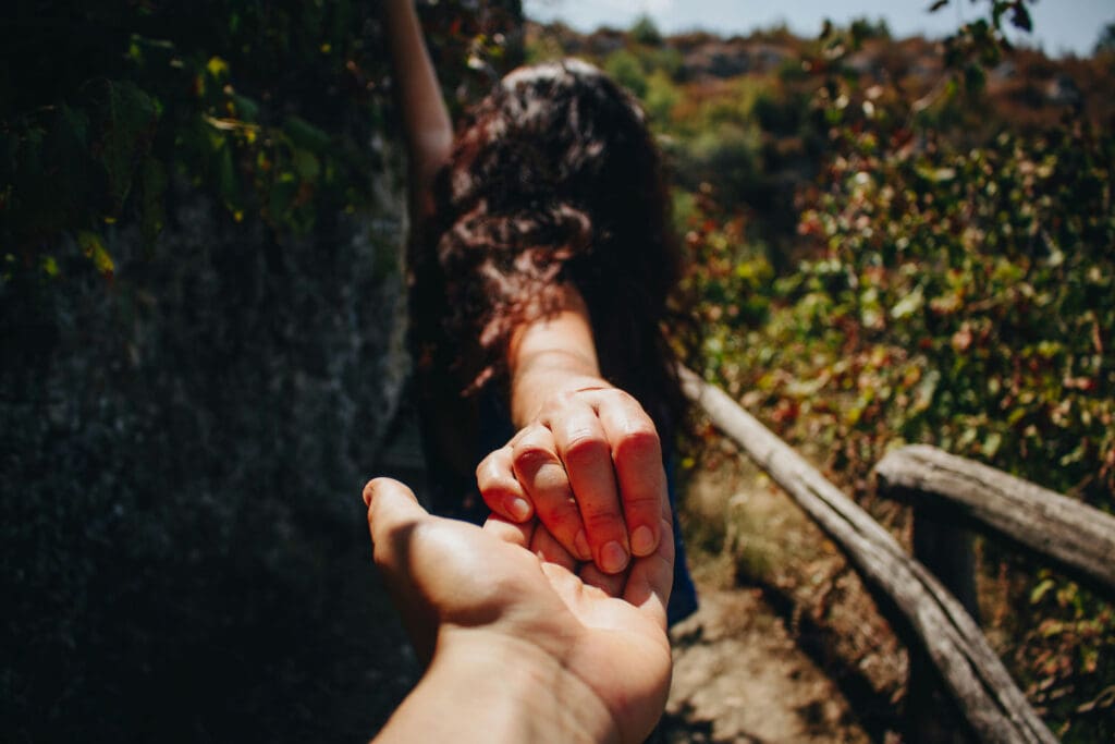 A woman with long hair walks along a rustic path, leading the viewer by the hand. The scene is surrounded by greenery and wooden railings, and the sun casts a warm glow on the landscape. Her back is turned to the camera.