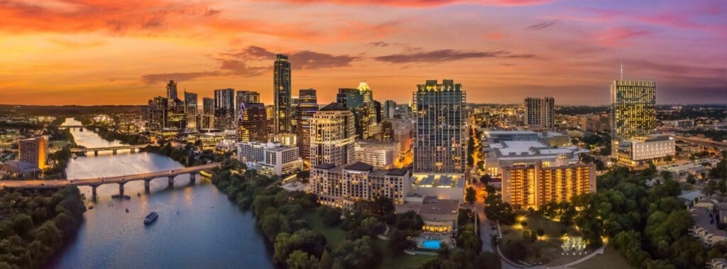 Panoramic view of a city skyline at sunset with a river in the foreground and illuminated buildings. The sky is vibrant with shades of orange, pink, and purple, reflecting on the water. A bridge spans the river, connecting parts of the city.