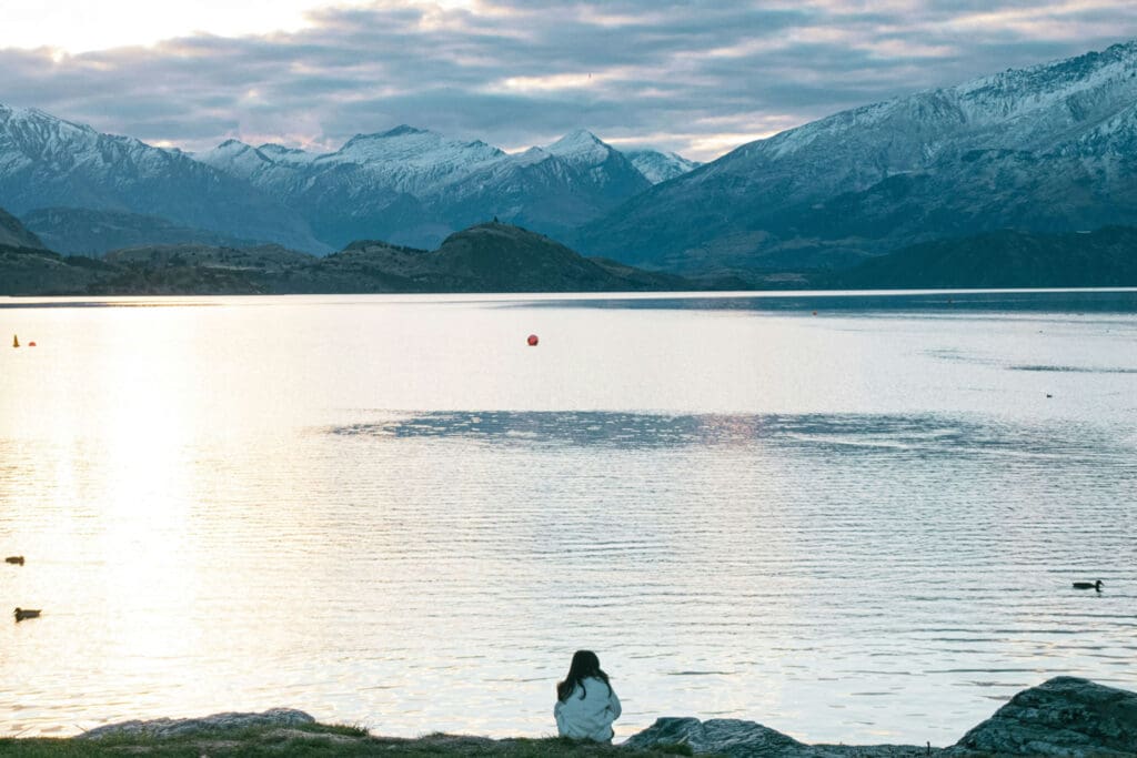 A person sits on a grassy shoreline, gazing at a tranquil lake surrounded by snow-capped mountains under a cloudy sky. Ducks swim nearby, and a buoy floats on the water reflecting soft sunlight.