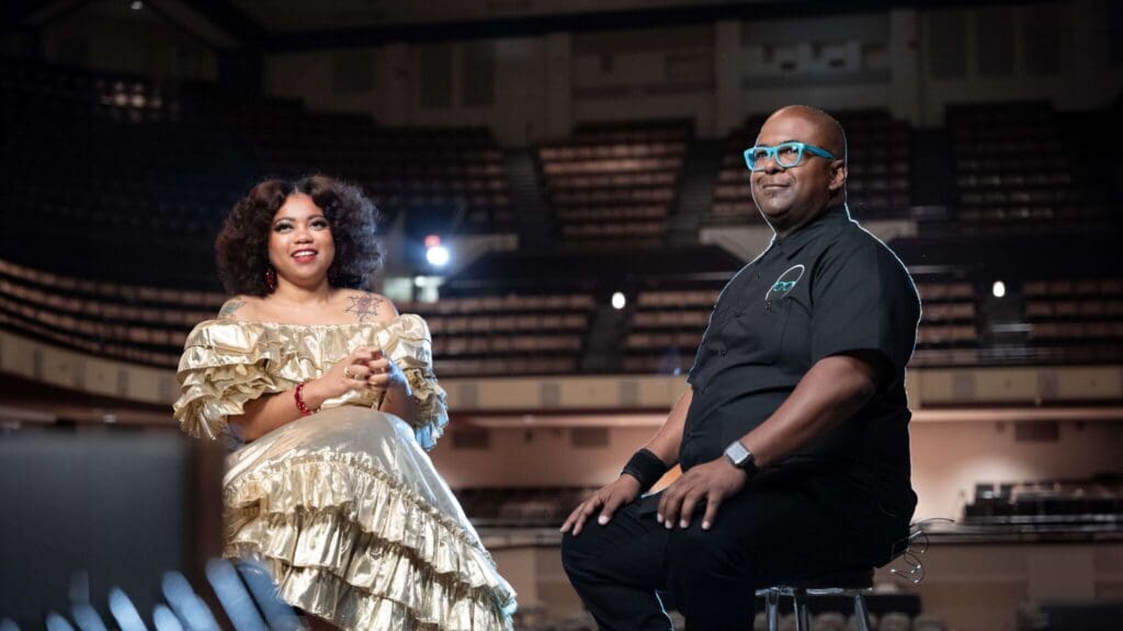 Two people are seated onstage in a well-lit auditorium. The woman on the left wears a gold ruffled dress, and the man on the right wears glasses and a black shirt. Theyre both smiling and appear mid-conversation.