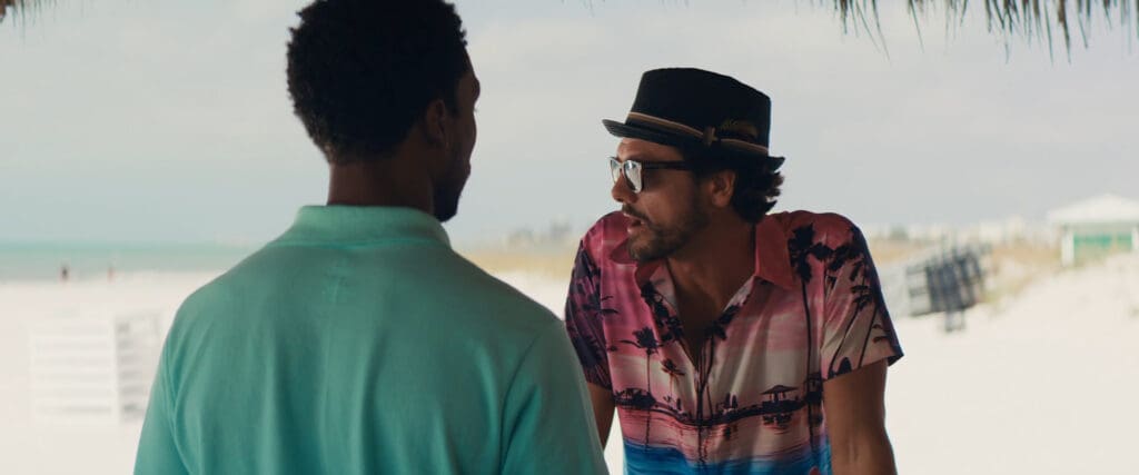 Two men are talking on a beach. One, in a turquoise shirt, faces away. The other, wearing a pink floral shirt, eyeglasses, and a hat, leans on a wooden surface. The background shows sand, a blue sky, and distant beach structures.