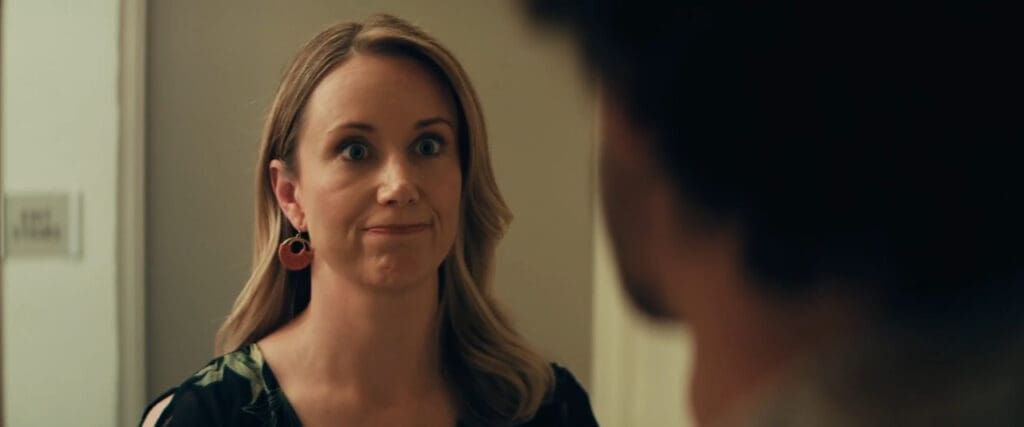 A woman with long blonde hair and pink earrings looks surprised or concerned. She is standing indoors, facing a person whose back is to the camera. The setting appears to be a home or office.