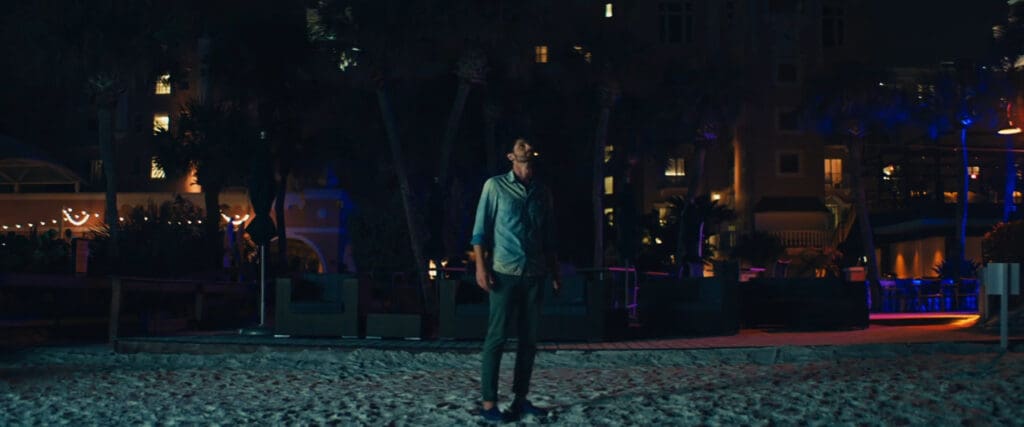 A man stands on a sandy beach at night, surrounded by dimly lit buildings and trees. The scene is dark, with colorful lights from windows and outdoor areas softly illuminating the background.