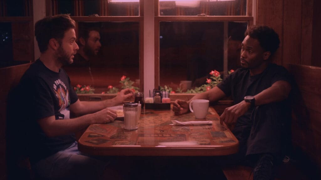 Two men sit across from each other in a dimly lit diner booth, engaged in conversation. One gestures with his hand, and the other listens with a cup of coffee in front of him. The table is decorated with various photos.
