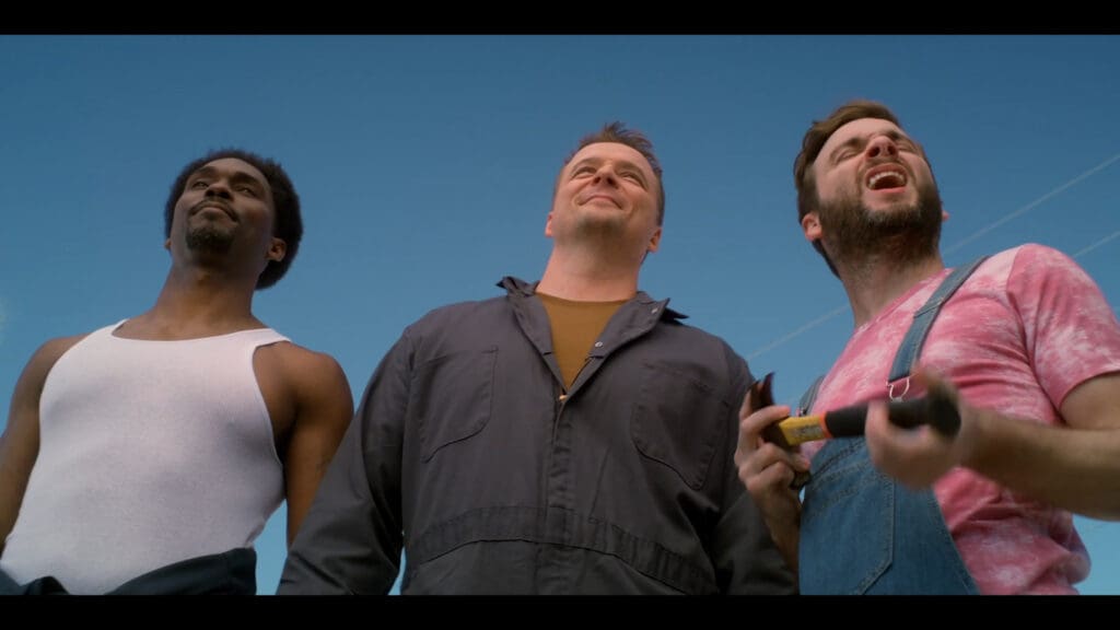 Three men smiling and looking up against a blue sky. The man on the left wears a white tank top, the middle man wears a dark jumpsuit, and the man on the right wears a pink shirt and denim overalls, holding a tool.