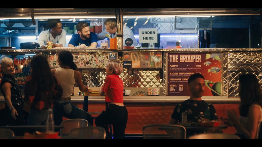 People sit at a counter of a food truck with a vibrant menu. Three men inside the truck chat, while customers outside enjoy their meals. A sign reads ORDER HERE. The scene is lively and colorful, with the trucks bright lights illuminating the area.