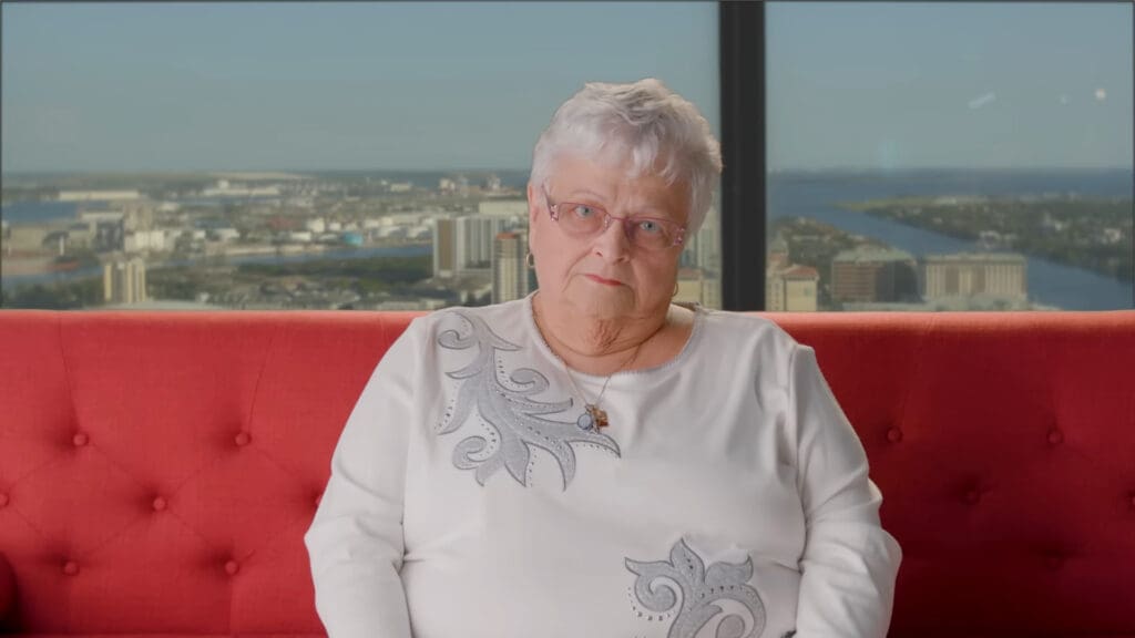 An elderly woman with short white hair and glasses sits on a red couch. Shes wearing a white sweater with decorative patterns and a pendant. A cityscape is visible through the large windows behind her.