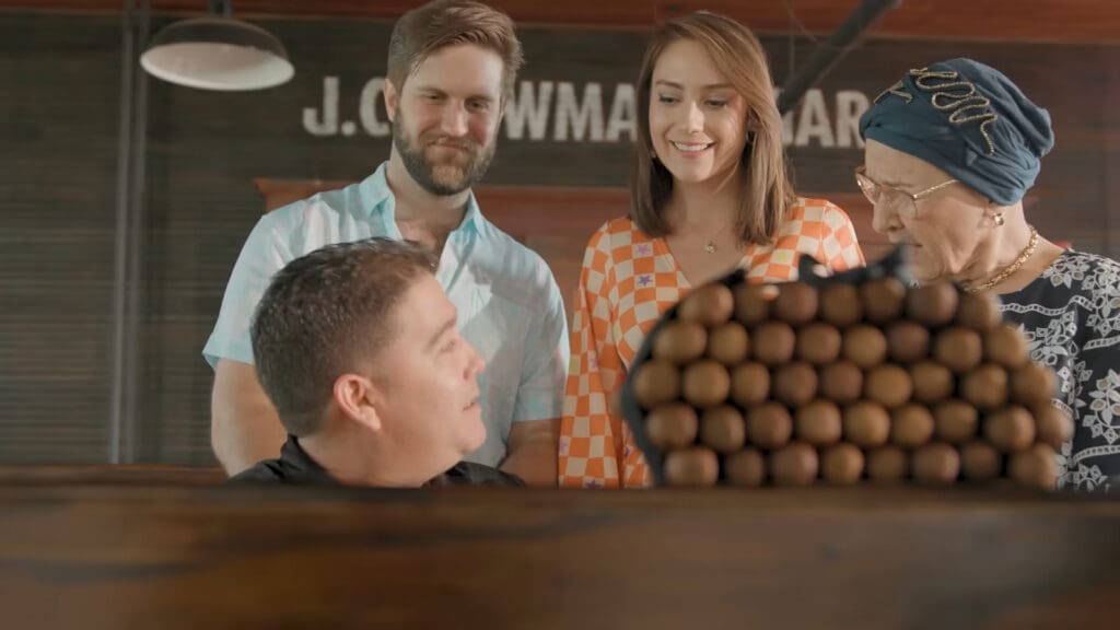 A group of four people is gathered around a wooden table looking at a stack of cigars. One person is seated facing the cigars, while the other three stand behind him, observing. The room has rustic decor with a sign in the background.