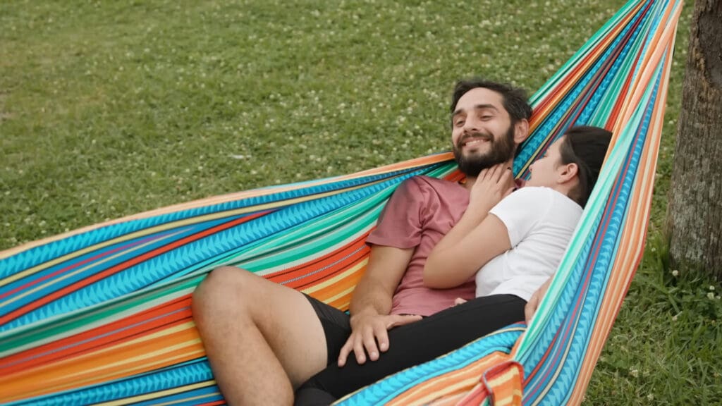 A couple relaxes in a colorful hammock outdoors. The man in a pink shirt smiles with his eyes closed, while the woman in a white shirt rests her head on his shoulder, looking content. Green grass is visible in the background.