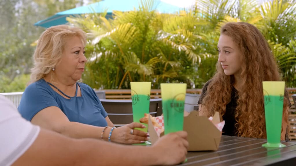 An older woman and a younger woman sit at an outdoor table, holding green drinks. They are engaged in conversation. There are plants and an umbrella in the background, and a takeout container is on the table.