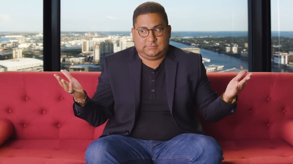 A person with glasses and short hair is sitting on a red sofa, shrugging with palms facing up. They are wearing a dark blazer and jeans. A large window offers a view of a cityscape and river in the background.