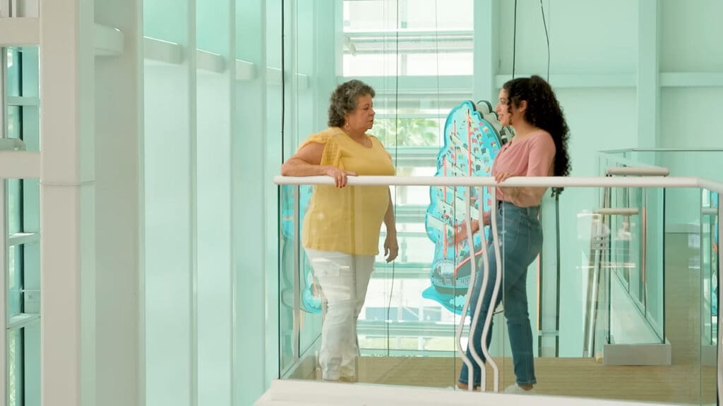 Two people stand on a modern glass balcony. One person, wearing a yellow shirt, faces another in a pink shirt and jeans. They are engaged in conversation, with a colorful butterfly artwork in the background.
