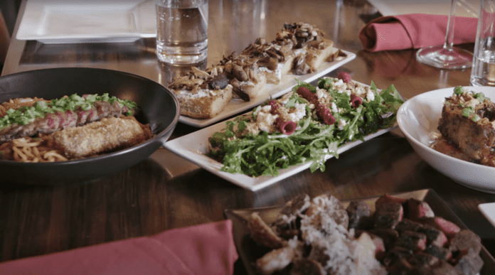 A table with a variety of dishes, including a bowl of pasta topped with meat, a rectangular plate of grilled mushrooms on toast, a salad with greens and meat, and a plate of steak slices. Red napkins and glassware are also visible.