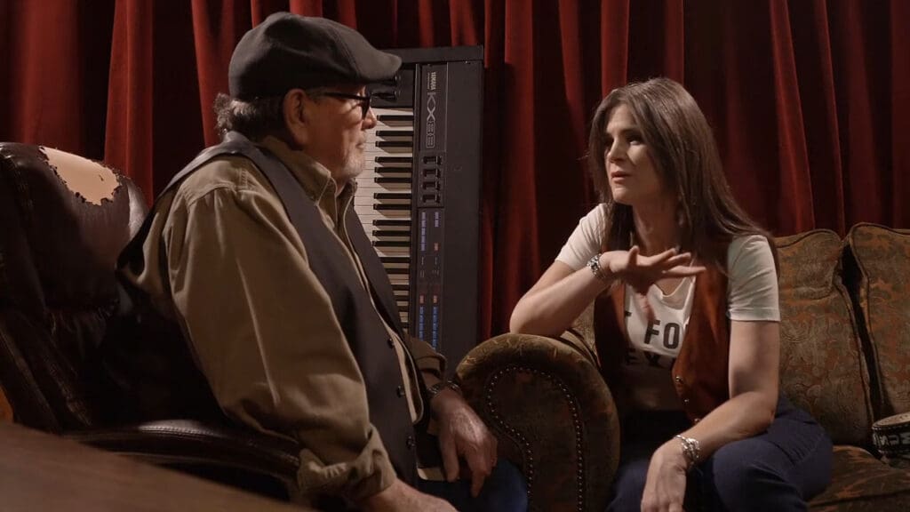 Two people sitting and talking in a cozy room. A man in a cap and vest sits on the left, while a woman in a white shirt and brown vest gestures while speaking on the right. A keyboard is visible in the background against red curtains.