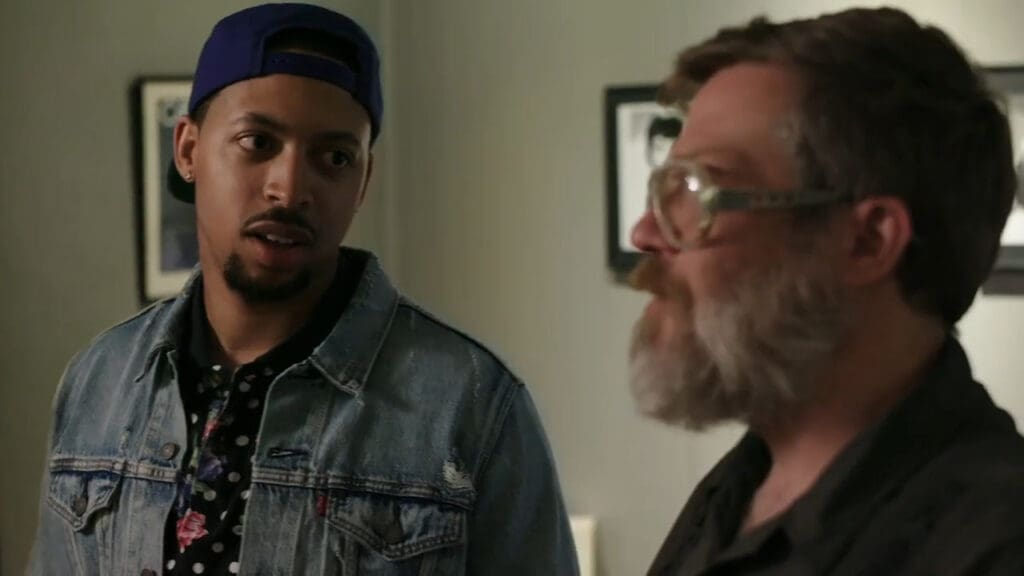 Two men are indoors. The man on the left wears a blue cap, denim jacket, and patterned shirt. The man on the right has glasses, a beard, and wears a dark shirt. They seem to be engaged in conversation. The background shows framed pictures on the wall.