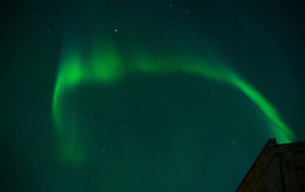 Green aurora borealis arcs across a starry night sky, creating a luminous glow. The bottom corner shows the silhouette of a structure.