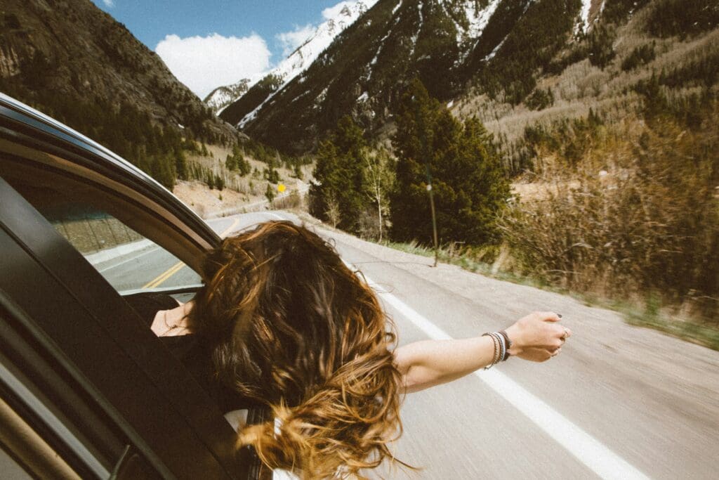 Person with long hair leaning out of a car window, hand outstretched. The car is driving on a winding road through a mountainous landscape with green trees and snow-capped peaks under a blue sky with scattered clouds.
