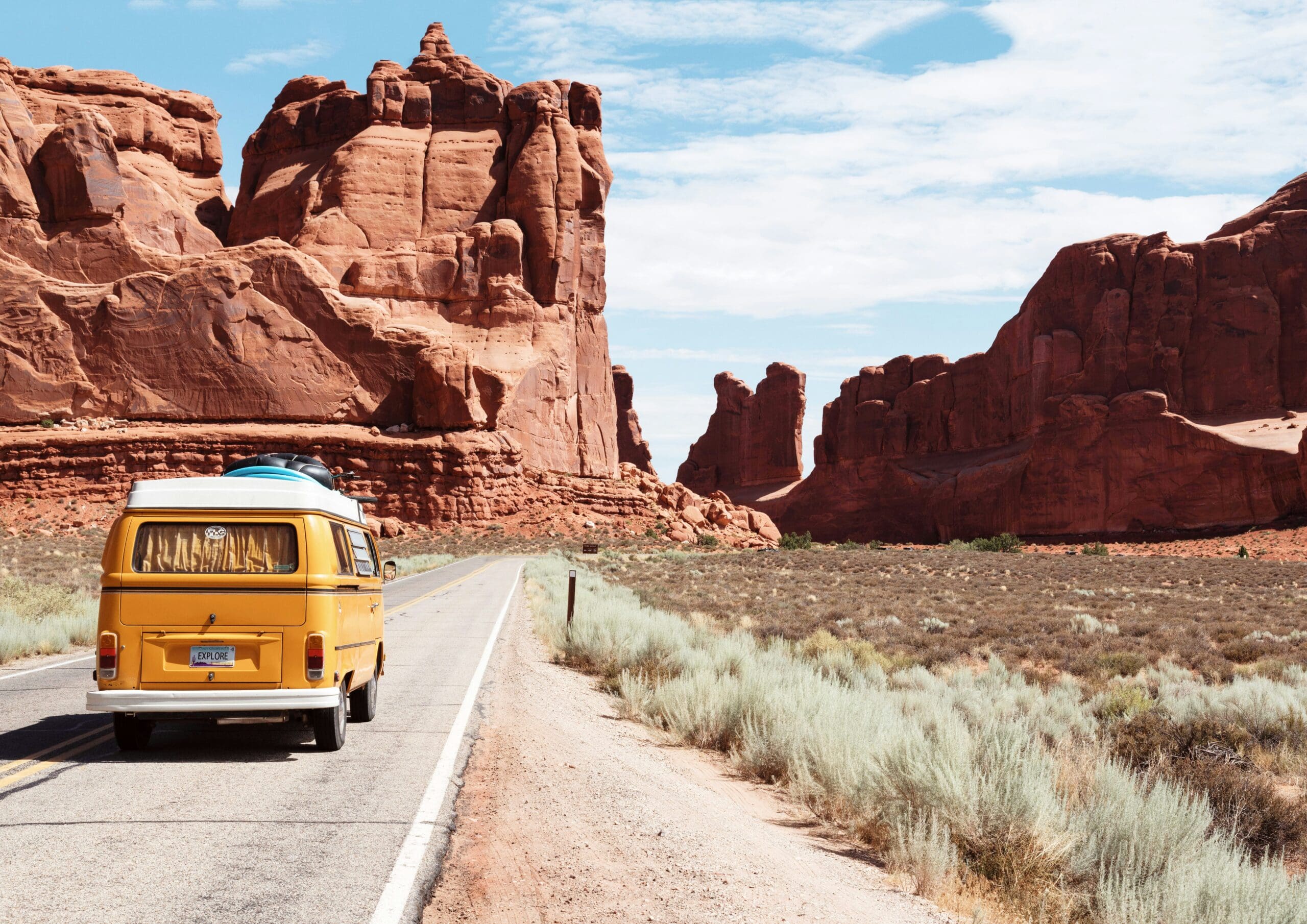 A yellow camper van with a surfboard on top drives down a road through a desert landscape with towering red rock formations under a blue sky with scattered clouds. Shrubs and dry grass line the roadside.