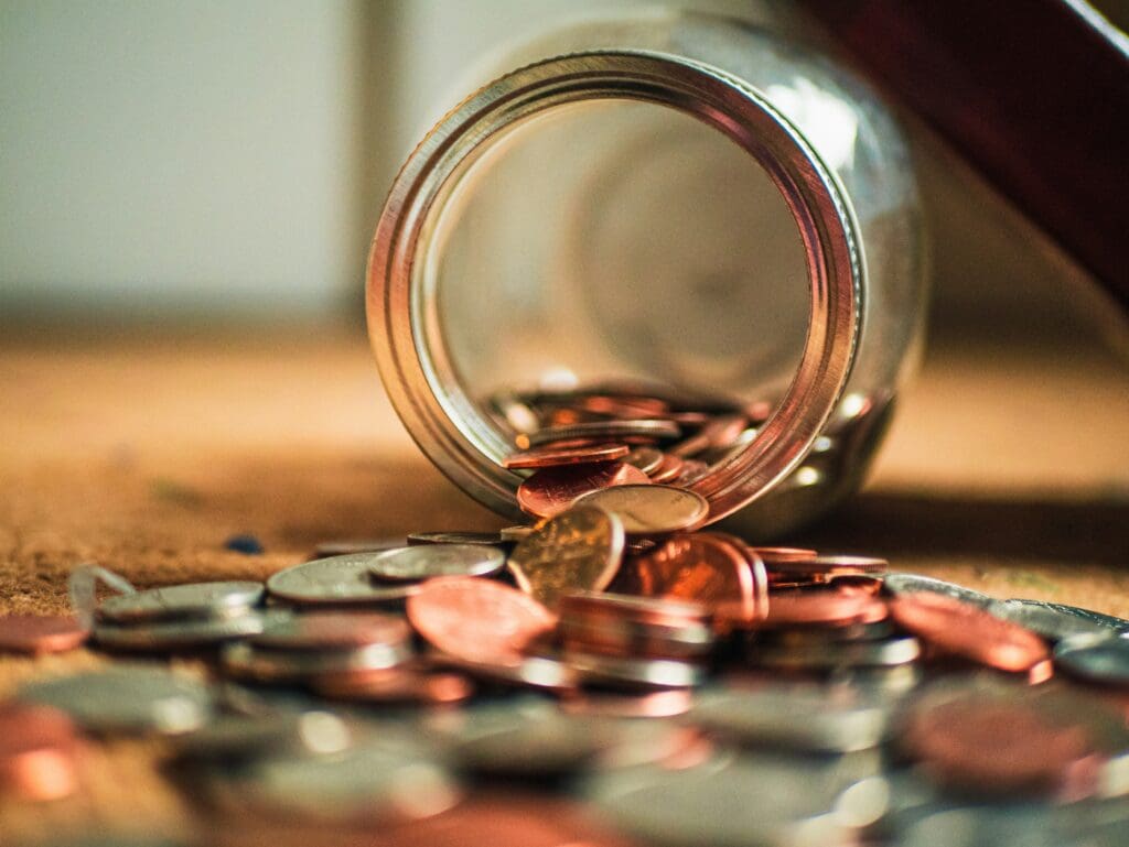 A glass jar tipped over with assorted coins spilling out onto a wooden surface. The coins are a mix of silver and copper colors, scattered around the open jar.