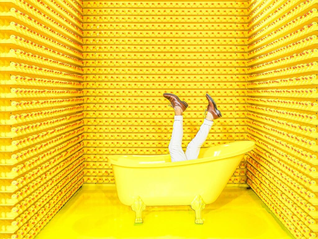 Person with legs up in the air, wearing white pants and brown shoes, lounging in a bright yellow bathtub. The room is filled with shelves lined with numerous yellow rubber ducks, creating a playful and whimsical atmosphere.