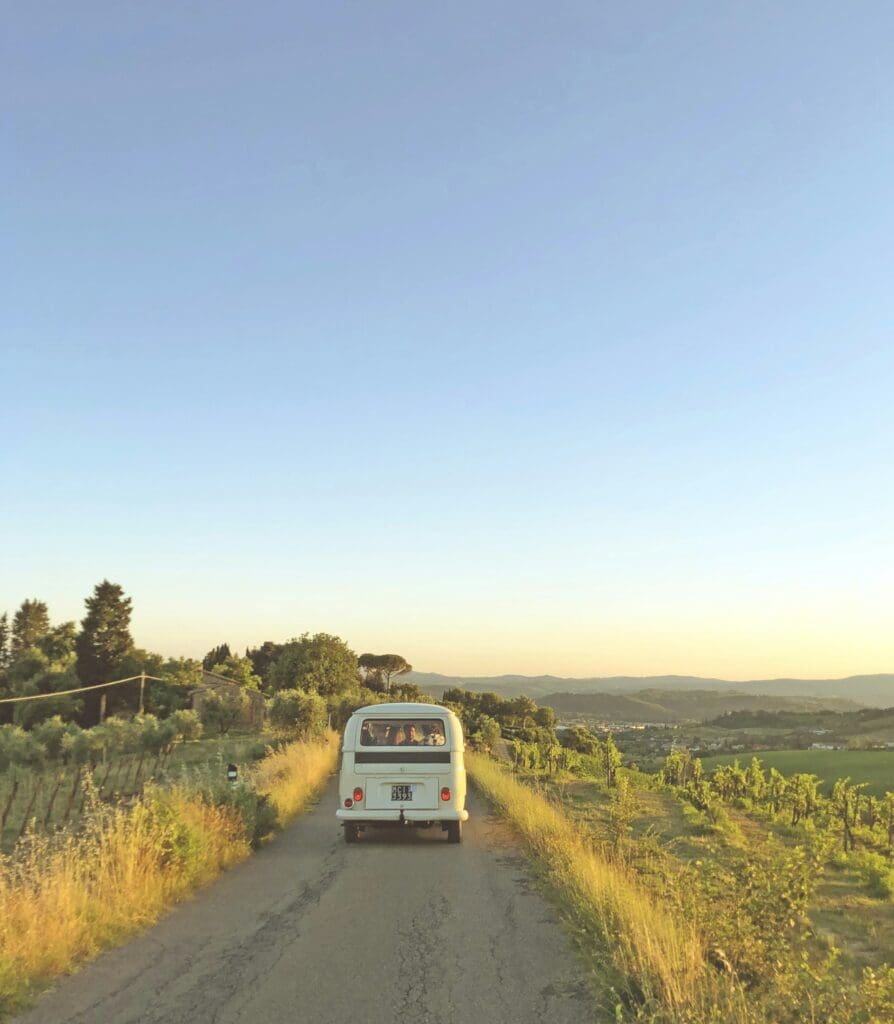 A vintage car drives down a narrow, rural road surrounded by fields and greenery. The sky is clear with a warm light, suggesting sunset. Trees and distant hills are visible on the horizon.