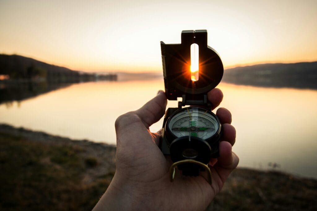 A hand holds a compass in the foreground with the sun setting behind it. The sun is aligned through the sight of the compass. Calm water and distant hills are in the background, with warm colors in the sky.