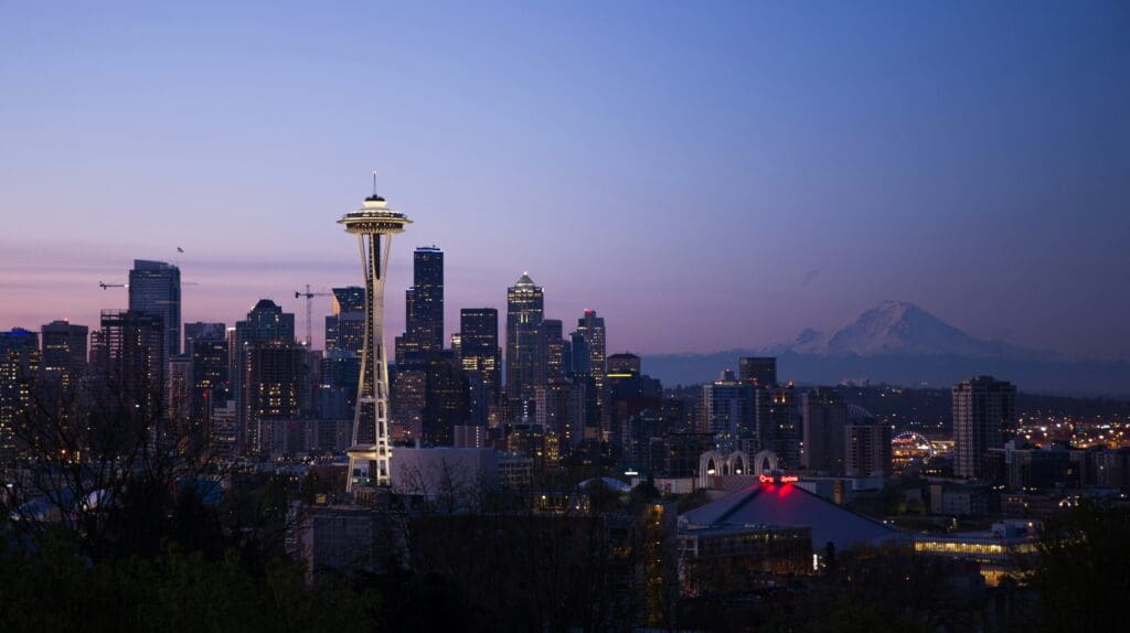 Seattle skyline at dusk with the Space Needle prominently visible. Skyscrapers are lit up, and Mount Rainier is in the background under a clear, gradually darkening sky.