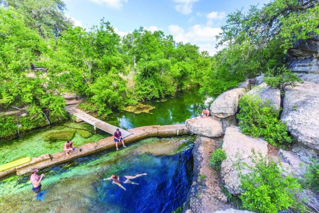 A scenic natural swimming area surrounded by lush green trees. Several people enjoy swimming and relaxing on the rocks near a clear blue pool of water. A small wooden bridge crosses the water, and a rocky ledge is visible to the right.