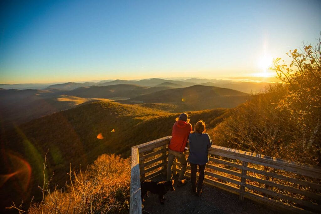 Two people and a dog stand on a wooden observation deck, overlooking a scenic view of rolling mountains bathed in golden sunlight during sunset. The sky is clear, enhancing the views vibrant colors.