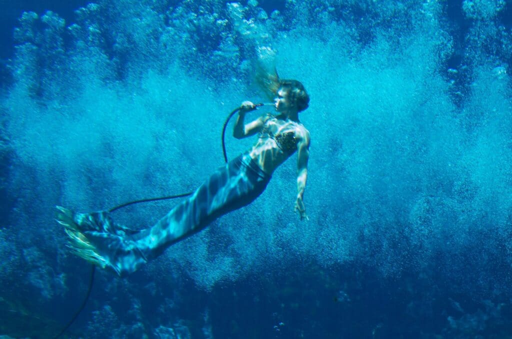 A person dressed as a mermaid is underwater, holding a long black hose. Bubbles surround them, creating an enchanting blue background. Light filters through the water, casting shadows and enhancing the dreamlike atmosphere.