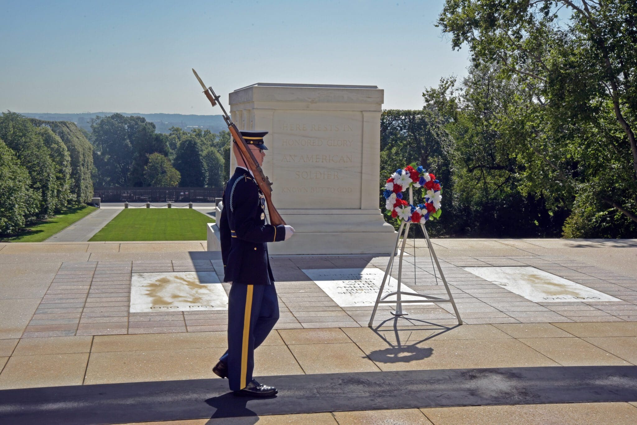 A soldier in dress uniform with a rifle on his shoulder walks in front of the Tomb of the Unknown Soldier. A floral wreath in red, white, and blue stands nearby. Trees and hills are visible in the background under a clear sky.