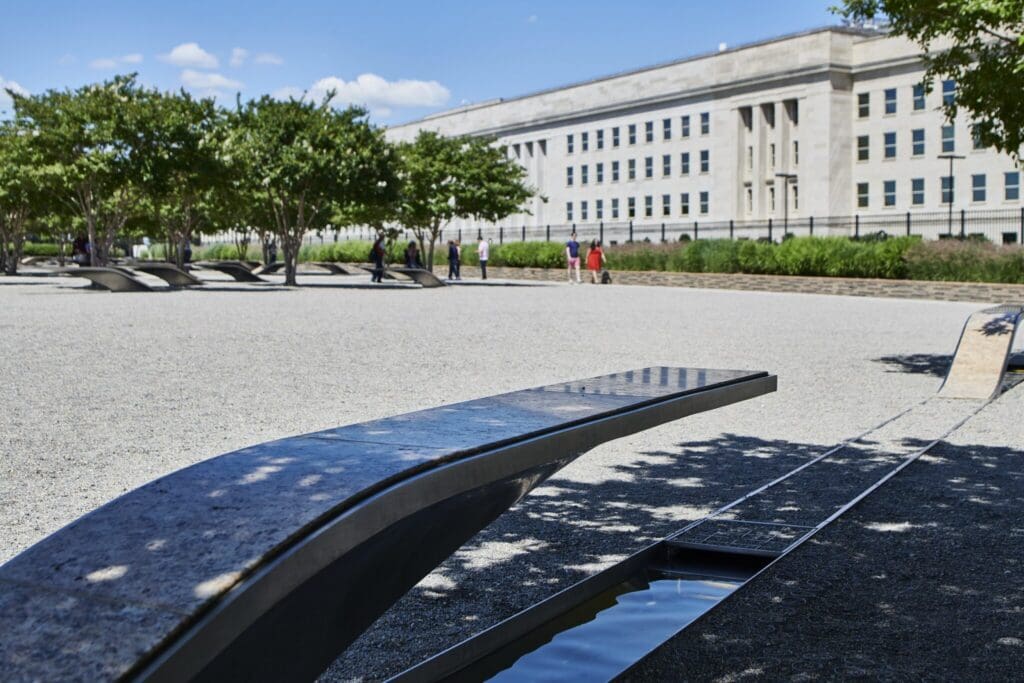 Memorial with curved benches over small reflecting pools, set in a gravel area with trees in the background. A large building is visible under a clear blue sky. People are walking in the distance.